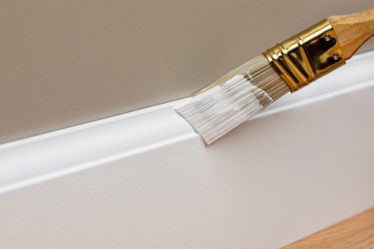 Paintbrush applying white paint to skirting board
